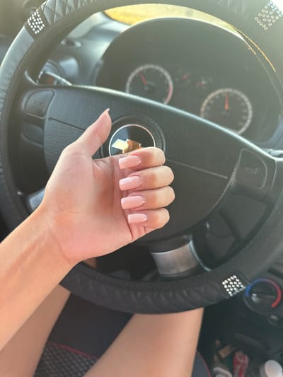 A first-person perspective shot from inside a car shows a person's hand with light pink squared-off fingernails resting on the steering wheel. The steering wheel has a black cover with diamond-shaped stitching and some sparkly embellishments. The car's dashboard is visible in the background, including the speedometer and other gauges, as well as the horn button on the steering wheel. The lighting suggests it is daytime, with sunlight streaming in. The location is not identifiable as Iordanovca, Moldova based on the image alone.