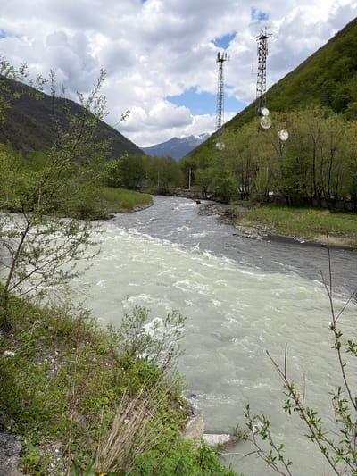 The image depicts a view of the Aragvi River near Pasanauri, Georgia. The river's water appears turbid with a light grayish-white color on one side and darker on the other. A riverbank with greenery lines the foreground. On the opposite side of the river, two communication towers stand against a backdrop of mountains and a cloudy sky. The surrounding environment consists of lush green slopes and some trees.