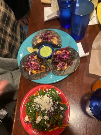 The image shows a table in a restaurant with three black tacos, a bowl of salad, two blue glasses, and a partly visible person wearing a plaid shirt. The tacos are on a blue plate and contain red cabbage, fish, and white sauce. The salad consists of leafy greens, grated cheese, red cabbage, and orange shavings, and is served in a red bowl. The glasses appear to be empty and are placed next to a white napkin on the table. The table appears to be made of wood. The location of this image is most likely Greenwich, United States.