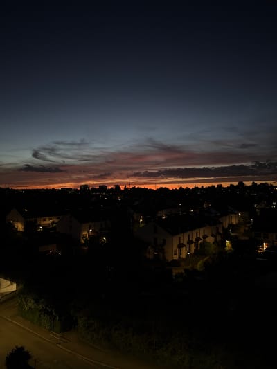 The image shows a twilight scene over a residential area in Sindelfingen, Germany. The sky is a deep gradient of blue and purple, transitioning to a warm orange glow near the horizon where the sun has set. Wispy clouds are illuminated by the setting sun, creating streaks of pink and orange across the sky. Below the sky, the silhouettes of buildings and trees create a dark landscape. Lights are visible in some of the windows of the houses, indicating that it is evening. The foreground is very dark, with only hints of a street and some greenery visible. The overall impression is one of a quiet, urban evening.