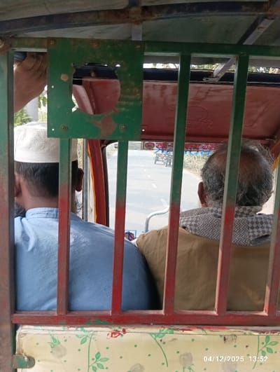 The image depicts a forward-facing view from inside a moving vehicle, likely a local transport, in Kaliganj, Bangladesh. Two adult males are seated in the vehicle's front passenger section, viewed from behind. The man on the left wears a light blue patterned shirt and a white skullcap, revealing dark hair. The man on the right, with gray curly hair, wears a plain brown garment and a checkered scarf around his neck. The interior structure of the vehicle features prominent, somewhat rusted, green vertical metal bars and a red horizontal bar. A dark green square metal plate with a circular cutout is affixed to the upper part of the framework. The seat in the foreground is upholstered with a light-colored cushion featuring a green leaf and red floral pattern. Beyond the vehicle's structure, a paved road extends into the distance under a bright sky, suggesting daytime. Another three-wheeled vehicle is visible on the road ahead. Trees and sparse vegetation line the road's edges. A faint sign with Bengali script is discernible in the mid-ground. A hand is visible in the upper left corner, grasping a bar. The bottom right corner displays a digital timestamp: "04/12/2025 13:52".