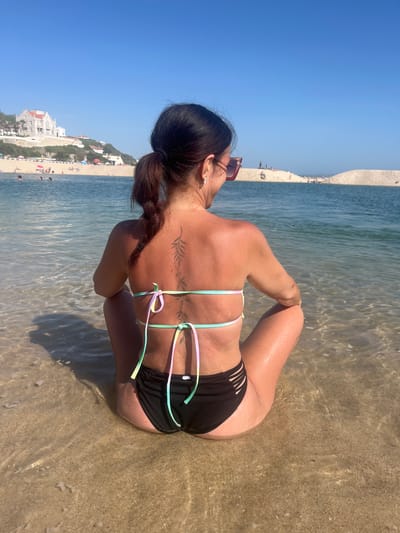 A woman sits on a sandy beach in the city of Caldas da Rainha, Portugal, with her back to the camera. She is wearing a black bikini with a pastel-colored halter top and sunglasses. Her hair is pulled back in a ponytail, and she has a tattoo on her back. The woman is facing the ocean, with a sandy beach and buildings visible in the background. The water is clear and shallow, with gentle waves lapping at the shore. The sky is a clear blue, and the sun is shining.