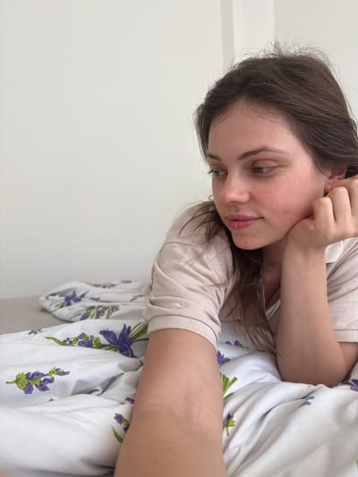 A young woman with dark hair is lying in a bed with white sheets patterned with purple lavender flowers. She is wearing a light beige shirt and has one hand near her ear. She is looking down and to the left. The wall behind her is white. The location is not discernible from the image. Therefore, the city of Kőröshegy, Hungary cannot be determined.