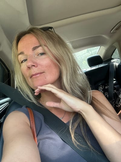A woman is sitting in the passenger seat of a car, facing the camera. She has blonde hair and is wearing a grey top and a seatbelt. Her left hand is resting on her chin. The car interior is visible, including the headrest of the seat behind her and a portion of the car's ceiling. Sunlight streams in from outside, illuminating her face and hair. The scene is likely outdoors, as indicated by the sunlight and glimpses of greenery through the car window. The exact location is not specified in the image, and therefore cannot be confirmed as being in the city of Rasskazovo, Russia.