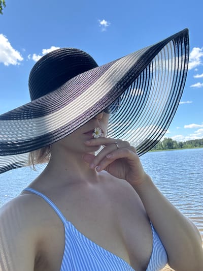 A person wearing a wide-brimmed black and white striped hat and a blue and white striped bikini top stands on a sandy shore, with a body of water and trees in the background. A daisy is held to the person's lips. The sky is blue with scattered clouds. The location is Lendži, Latvia.