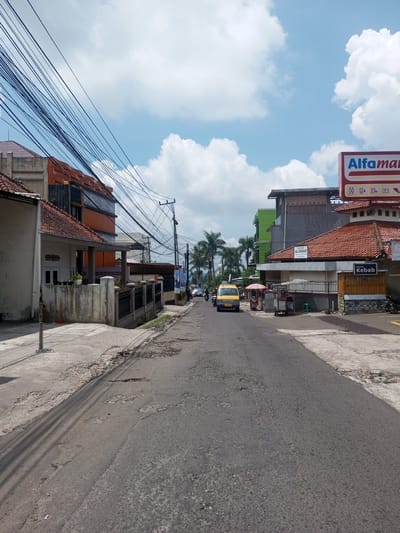The image depicts a street in a seemingly Southeast Asian location. The road is paved but shows signs of wear and tear with multiple potholes. On either side of the street are buildings, a mix of residential and commercial structures, including a shop with a sign that reads "Alfaman" and a "Kebab" stall. Utility lines are strung haphazardly across the sky above the street. A small yellow van is visible driving along the street, and there are also some scooters parked on the side. The sky is blue with scattered white clouds. The location is likely not in Xayabury District, Laos as it's written in Indonesian, the "Alfaman" sign is an Indonesian convenience store, and the architecture does not match.