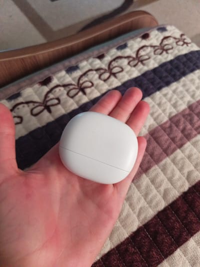 A person's hand holds a small, white, oval-shaped object, possibly an earbud case, in the center of the frame. The hand is cupped, with fingers curled around the object, and the palm is visible. The object has a subtle seam running across its surface, suggesting it might be a two-part enclosure.
The background is a textured surface, appearing to be a quilt or blanket. It features a pattern of alternating colored stripes, primarily shades of beige, purple, and dark red, with a quilted diamond pattern within the lighter sections. Interspersed among the lighter sections are decorative elements that resemble stylized bows or floral motifs in a dark brown color. A wooden armrest of a chair is visible in the upper left corner, hinting at a domestic or indoor setting. The lighting suggests it might be daytime, possibly indoors with natural light, as there are no harsh shadows. The overall mood appears calm and casual. No visible text is present in the image. The location is identified as Sukhodol, Russia.