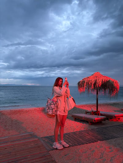 A woman stands on a beach in Alimos, Greece, taking a photo with her smartphone. She is wearing a white outfit and sneakers, and carries two bags. To her right are beach umbrellas, tables, and chairs, bathed in red light. The beach is a mix of sand and a wooden platform. The sea is visible behind her. Above, the sky is filled with dark, cloudy formations.