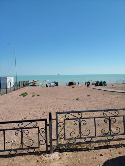 The image depicts a sunny day at a beach in Toranghilik, Kazakhstan. The foreground features a black wrought-iron fence with decorative swirls. Beyond the fence, a sandy beach is visible, with several tents set up. A paved pathway leads towards the water. In the distance, the turquoise water of the sea is calm, with a pier extending into it. Several people are seen swimming and relaxing in the water. Further out, a boat with people on it and a parasailer are visible. A white building with a fence around it is situated on the left side of the image, and a street lamp stands nearby. The sky is clear blue.