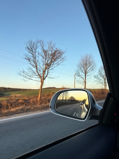 The image displays a view from inside a moving vehicle on a rural road near Kamburovo, Bulgaria, during the late afternoon. The vehicle's silver-grey side mirror is prominently positioned on the right, reflecting the road behind. Within the mirror's reflection, a two-lane asphalt road recedes into the distance, bordered by bare deciduous trees, with a white sedan observed far down the road against a yellowish-orange sky. Outside the vehicle, to the left of the mirror, the asphalt road is visible. The adjacent landscape includes bare deciduous trees, utility poles supporting power lines, and fields with green and brown vegetation, leading to distant hills under a clear blue sky that brightens to yellow at the horizon. A dark interior car door panel with a small red indicator light is visible in the lower right portion of the frame. No individuals are depicted.
