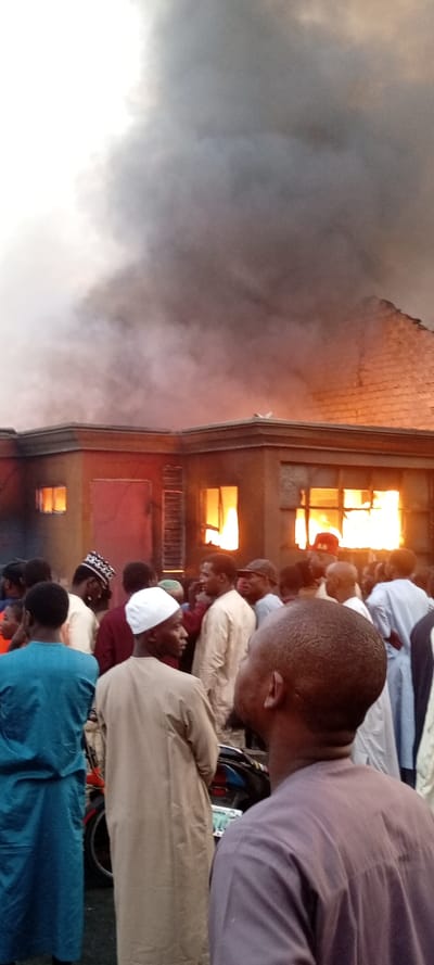 A dramatic and somber scene unfolds in this vertical-format image, capturing a building engulfed in flames with a large crowd of onlookers. The focal point is a low-rise building, possibly a single-story structure, from which intense orange and yellow flames are actively bursting through multiple windows. At least four windows on the front and right side of the building show visible fire, indicating a significant internal blaze. The building's walls appear to be a light tan or brown color, with sections above and around the windows visibly blackened and discolored by smoke and heat.
Above the burning building, a colossal plume of thick, dark gray and black smoke billows upwards, dominating the upper two-thirds of the image. The smoke is so dense that it largely obscures the sky, creating a somber, ominous backdrop. The light quality, with the sky appearing brighter in the top left corner but heavily overcast by smoke elsewhere, suggests the event might be occurring during late afternoon or early evening.
In the foreground and midground, a substantial crowd of people, predominantly men, are gathered. They are positioned with their backs mostly to the camera or facing slightly to the side, all looking towards the burning structure. Many individuals are dressed in traditional attire, including long, flowing robes in various shades of brown, tan, blue, and purple. Several men wear head coverings such as kufis or prayer caps, one prominently white, another with a black and white pattern. A man in the immediate foreground, with his back partially to the camera, wears a dark purple garment, while another on the far left wears a vibrant blue robe. Their presence indicates a public response to the incident, likely observing the fire. A motorcycle or scooter is partially visible behind some of the onlookers in the lower middle portion of the image, suggesting an urban or semi-urban setting. No discernible text is legible anywhere in the image, including on clothing or the partially visible vehicle. The overall impression is one of an unfolding emergency, drawing a concerned or curious crowd.