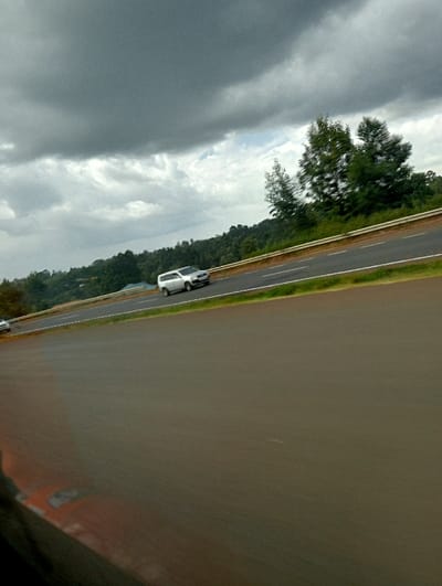 A grey SUV drives along a highway under a cloudy sky. The road curves slightly, with green vegetation and trees visible on the roadside. The image appears to be taken from a moving vehicle, focusing on the road and the car in motion. The overall scene suggests a drive through a rural or suburban area on an overcast day.