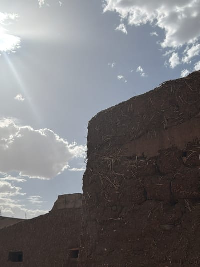 An upward-angled view reveals part of an earthen structure against a partially cloudy sky, situated in Sidi Bou Othmane, Morocco. The structure's rough texture, interspersed with straw, suggests traditional building techniques. Sunlight streams from behind a cloud, creating a bright flare in the sky. A section of another earthen building with possible window openings appears partially in the lower right. There are no people visible. The environment suggests a rural setting.
