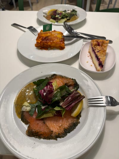 A close-up, overhead shot shows three plates of food arranged on a white table. In the foreground, a white plate holds a generous portion of smoked salmon, topped with fresh greens and a lemon wedge, accompanied by a creamy yellow dressing and a fork. To the upper left, a white plate displays a slice of lasagna adorned with a small green flag. To the right, a small white dish contains a slice of blueberry crumble cheesecake. Cutlery, including a fork and spoon, is visible between the lasagna and cheesecake. The background features out-of-focus green chairs and a glimpse of a white surface. The setting appears to be a restaurant or cafeteria, likely within the city of Goyang-si, South Korea, given the context provided.
