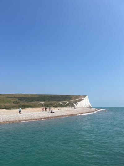 This is a photograph of the Seven Sisters cliffs in Seaford, United Kingdom, on a clear, sunny day. The image is taken from a low angle, looking towards the sea. In the foreground, the turquoise water is calm with small waves lapping at the shore. The beach is made of pebbles, and a few people are visible walking along it or sitting on the shore. Further back, the iconic white chalk cliffs rise dramatically, meeting a green, rolling hillside. The sky above is a vast expanse of clear blue. The scene captures a typical summer day at this popular coastal location.