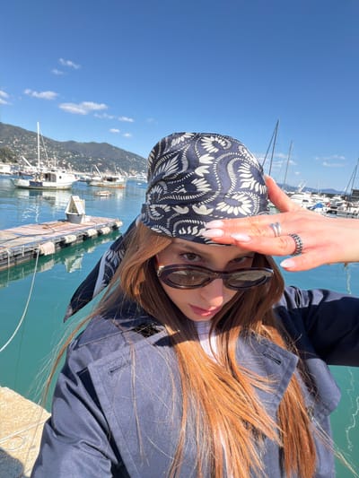 A woman with long auburn hair, sunglasses, and a patterned headscarf is photographed outdoors on a sunny day. She is wearing a dark blue jacket. Her hand, adorned with rings, is positioned above her head. The background reveals a harbor scene in Santa Margherita Ligure, Italy, with boats, docks, buildings along the coastline, and a clear blue sky with scattered clouds. The water in the harbor appears greenish-blue.