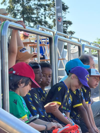 A group of children and adults are riding on an open-top tour bus in Fentange, Luxembourg. The bus is equipped with a chrome safety railing system. The riders are gazing upwards, presumably at a point of interest. The children are wearing colorful baseball caps and soccer jerseys, suggesting a team outing. The backdrop includes trees and a street sign.