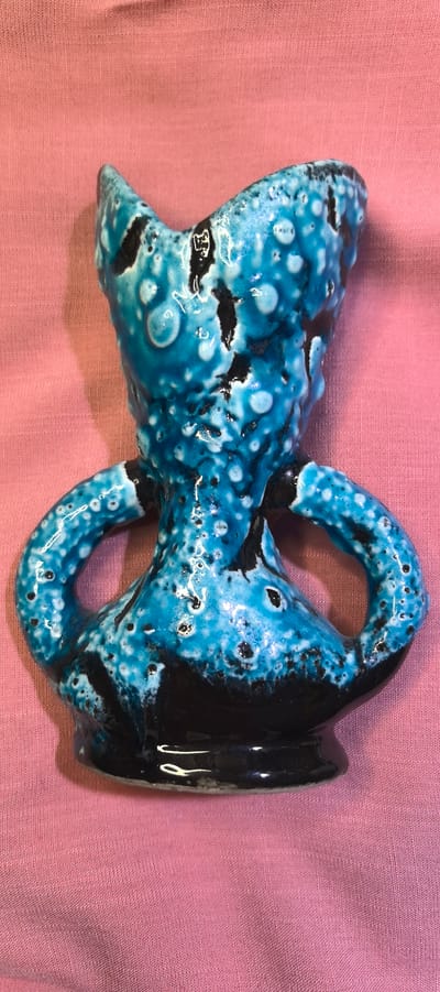 A blue and black ceramic vase with two handles is displayed on a pink fabric surface. The vase has a textured, bubbled glaze with dark specks. The fabric is wrinkled and shows some slight staining. The image was taken in Villeneuve-le-Roi, France.