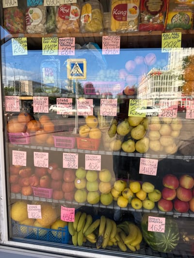 This is a close-up, eye-level shot of a fruit and dried fruit stall displayed behind a glass window, likely in an outdoor market. The scene is brightly lit, suggesting daytime. The reflection on the glass shows buildings and trees, hinting at an urban or town setting. The vendor is not visible, but the display is neatly arranged with various fruits and packaged dried goods.
Prominently featured are piles of apples in shades of red and yellow, bananas, plums, and a large watermelon. Several labels with prices and product names in Russian are visible, such as "ЯБЛОКИ" (apples), "БАНАНЫ" (bananas), "АРБУЗ" (watermelon), and "ГРУША" (pear). The prices are also in Russian currency. There are also packaged dried fruits and nuts with branding like "Kong" visible. A pedestrian crossing sign is reflected in the glass, further suggesting a public street location. The overall impression is one of a vibrant and well-stocked market stall.
