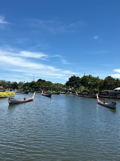The image depicts a waterway in Bang Pu Subdistrict Municipality, Thailand, with several longboats docked along the bank. The sky is a bright blue with wispy clouds. The water has a rippled surface. Trees line the bank, and a structure is visible behind them. A tall spire-like structure can be seen in the distance behind the trees.