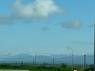 The image depicts a distant view of snow-capped mountains under a partly cloudy blue sky, likely taken from an elevated position within or near the city of Dzhiqumyr, Abkhazia. In the foreground, a green metal fence runs horizontally across the frame, with posts spaced at intervals. Beyond the fence, lush green grass and vegetation are visible. Several streetlights, characterized by their curved poles and dual lamps, are positioned along the fence line. Power lines are strung between poles, creating a network of wires above the vegetation. The mountains in the background are hazy blue, with patches of white snow on their peaks. The overall scene suggests a rural or semi-rural environment on a clear day.