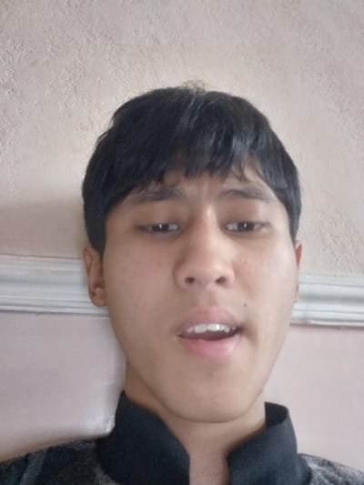 The image depicts a close-up, head-and-shoulders portrait of a young male, likely in his late teens or early twenties, against an indoor backdrop in Bagat, Uzbekistan. The subject has short, dark hair and light-to-medium brown skin. He is wearing a dark, possibly black, collared shirt. His eyes are directed downwards, and his mouth is slightly open, revealing both upper and lower teeth, suggesting a moment of speech or an unvoiced utterance. The background consists of a textured, light-beige or off-white wall. A white horizontal molding or trim is visible on the left side of the frame, behind the subject's head. The lighting is soft and even.