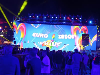 The image depicts a nighttime scene at an outdoor event, likely a Pride festival in Playa Del Inglés, Spain, based on the 'ASPA Pride Festival' signage displayed prominently on a large screen in the background. The screen features rainbow-colored hearts and rays. Above the screen, a rigging structure supports lights emitting yellow, red, and blue beams. A dense crowd of people fills the foreground, with individuals varying in dress and posture. The overall lighting is dark with a blue tint.