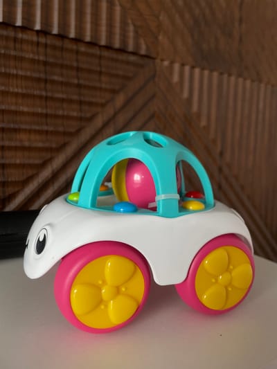 The image shows a colorful baby toy with a white, animal-shaped body and two pink wheels with yellow centers. A teal cage-like structure surrounds a ball filled with various colored balls. The toy is resting on a brown, woven surface and part of a white wall. It is not possible to identify any specific details relating to Средний Постол, Russia from the image.