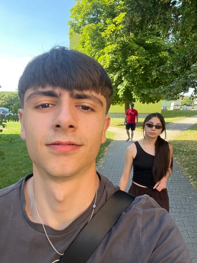 A selfie taken outdoors shows a young man in the foreground, his face close to the camera. He has dark, short hair and is wearing a dark grey t-shirt with a silver chain necklace. Behind him, a young woman with long, dark hair and sunglasses is looking towards the camera. She is wearing a black crop top and dark shorts. Further in the background, a person in a red t-shirt and blue shorts is walking away from the camera. The setting appears to be a park or a paved path alongside a grassy area with trees. The sky is blue and clear, suggesting it is daytime and the weather is pleasant. The scene is likely Dobřany, Czechia, as indicated by the location context.
