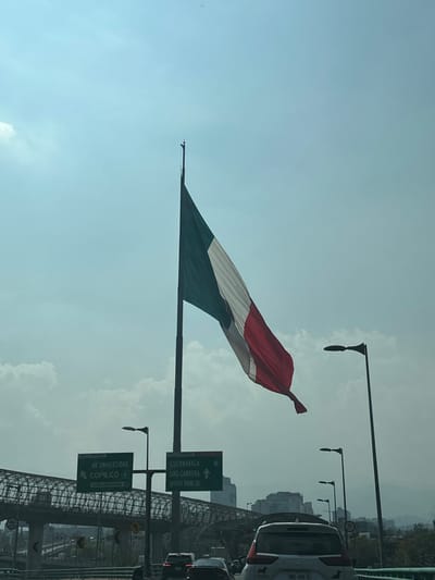 A large Mexican flag waves against a bright, cloudy sky. The flag's green, white, and red stripes are visible. Traffic and buildings line the lower right. Signage for "Cuernavaca, Luis Cabrera" and "Av. Universidad, Copilco" is prominently displayed. Several street lamps extend into the frame from the right. The image appears to be taken from a moving vehicle.