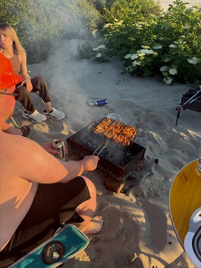 A group of people are having a barbecue on a sandy beach, likely in Povarovka, Russia, given the context provided. The scene is illuminated by the warm, late afternoon sun. In the foreground, a shirtless man, wearing black shorts and a hard hat, is seated and holding a can of beer. He appears to be tending to a grill filled with skewers of meat. To his left, a woman with blonde hair is seated in a chair, looking off to the side and eating something. Her legs are crossed, and she is wearing a brown tank top and dark pants. White Crocs are visible on her feet. In the background, there are bushes with white flowers and trees. The sand is marked with footprints and shadows. A blue spray can lies on the sand near the woman. Another smaller grill with tongs is visible further in the background. A yellow table with a bottle on it is in the right foreground.