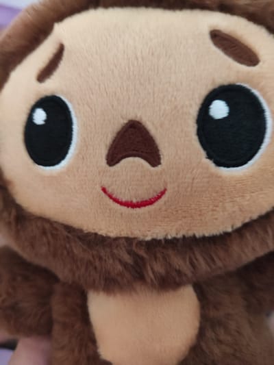 This media file is a close-up, vertically oriented photograph of a plush toy monkey. The subject is predominantly the monkey's face, which is made of light brown, fuzzy material. The monkey has large, black embroidered eyes with white highlights, giving it a somewhat surprised or wide-eyed expression. A small, dark brown embroidered nose is visible above a thin, red stitched smile. The outer edges of the image show the monkey's brown, furry arms and head. The background is out of focus but appears to be a textured surface, possibly fabric or a blanket, with hints of purple. The lighting suggests an indoor environment, possibly during the daytime, but no specific time or weather cues are evident. There is no visible text in the image. The setting is not specified beyond it being a photograph of a toy. No activity or event is taking place. The emotions conveyed are limited to the facial expression of the toy.