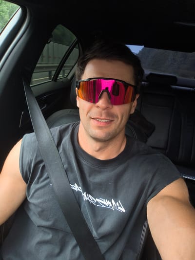 A man with short, dark hair is captured in a selfie from inside a car. He is wearing a dark gray, sleeveless t-shirt with white graffiti-style lettering on the front, and a black seatbelt is visible across his chest. He is also wearing distinctive oversized sports sunglasses with mirrored pink and orange lenses. The man is smiling, with his mouth slightly open, revealing his teeth. The interior of the car is dark, with black leather seats and a dark headliner. Outside the car windows, trees and foliage are visible, suggesting a setting in a more rural or suburban area, potentially along a road or highway. The backdrop appears to be a natural environment. The image does not provide any specific details indicating a location in Myasnoi Bor, Russia.