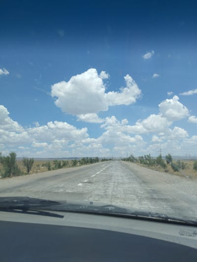 The image shows a road stretching into the distance under a bright blue sky with scattered white clouds. The road is paved and appears to be in fair condition, with visible white lane markings. To the sides of the road, there is sparse vegetation consisting of dry grasses and small, bushy trees. In the background, there are signs of human infrastructure, including power lines and possibly some distant buildings or structures. The perspective is from inside a vehicle, with the windshield and part of the dashboard visible in the foreground. There are no people or animals in the scene. The environment suggests a dry, possibly semi-arid region. While the image itself does not explicitly confirm the location as Besapan, Uzbekistan, the description aligns with potential landscapes in that region.