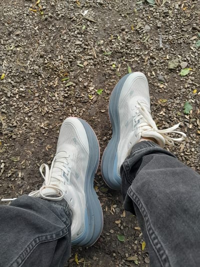 In Atabe, Georgia, a pair of white and gray athletic shoes are worn by a person sitting on a ground covered with small pebbles, dirt, and scattered leaves. The shoes have white laces and thick, layered soles with a gray and light blue hue, and a subtle pink accent along the bottom edge. The person is wearing dark gray denim jeans. The scene is outdoors, likely in a park or on a trail, indicated by the natural ground cover.