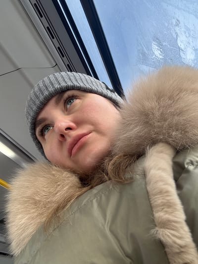 A person wearing a gray knit hat and a large fur-trimmed coat looks toward the camera. The individual's eyes are a striking green or gray. A window showing a cloudy sky is visible in the top left of the frame, suggesting an indoor setting. The perspective is slightly from below, looking up at the person. The image is rotated, so the person's head appears to be tilted.