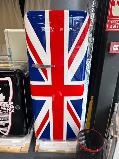 The image captures a Smeg appliance, likely a refrigerator or freezer, designed with the Union Jack flag prominently displayed across its front. The flag features bold red, white, and blue colors, with the brand name "SMEG" vertically embossed in silver lettering on the blue section on the left. A sleek, chrome handle extends horizontally from the red cross of the flag.
The appliance is situated within what appears to be a storage area, warehouse, or retail backroom, indicated by its packaging in white styrofoam inserts and wooden crate elements visible on the right. Below this appliance, another black Smeg unit is partially visible, also packaged. This lower appliance features white and red racing-themed graphics, including the text "24 HEURES DU MANS" and a checkered flag pattern, suggesting a motorsports design.
Above the Union Jack appliance, a dark metal beam or shelf runs horizontally across the frame. Draped over the top edge of the appliance and below the beam are tangled strands of clear string lights. On the wall above the beam, a red sign is partially visible, displaying white text that reads "POLSA D'ALARM," likely a truncated "PULSAR D'ALARMA" (Push Alarm in Catalan), consistent with the Andorra la Vella location. Next to this sign is a red fire alarm call point with a white symbol of two arrows pointing downwards to a button. To the far right, a black mesh cylinder, possibly a speaker or ventilation component with a red rim, is visible. Various pipes and hoses are also discernible in the upper right background. The scene suggests the appliances are new, in transit, or awaiting display/sale.