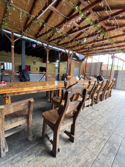 The image depicts a long wooden table with attached wooden chairs, likely in a restaurant or outdoor seating area in Картмазово, Russia. The table and chairs appear rustic and handcrafted. Above the table, there is a wooden structure with string lights and decorative garlands. The flooring is made of wood planks. Some personal belongings like bags are placed on the chairs. The background reveals the interior of a building with windows and other architectural features.