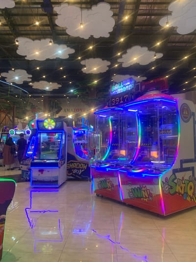 This image depicts a lively indoor entertainment center, likely an arcade or family fun zone, in Samarinda, Indonesia, illuminated by numerous vibrant lights, suggesting it's nighttime or a low-light setting. The scene is dominated by several brightly lit arcade machines with colorful LED accents.

In the center, a large claw or prize redemption game is prominent, featuring a clear casing and a cascade of rainbow-colored and blue LED lights. On this machine, the words "BALL MANIA" and the number "3917" are visible, along with "ulti-bal" and "CREDITS." Another adjacent machine prominently displays "WIN."

Further into the background, other arcade games can be seen, including one labeled "SHADOW BOY" and "AMAZONE," glowing with blue lights. A few indistinct figures of people are visible in the background, appearing to be enjoying the games, with one person in a brown skirt near a machine displaying "PEARL."

The walls are decorated with multiple white, cloud-shaped cutouts, illuminated by warm, glowing string lights, creating a whimsical atmosphere. The floor is a reflective, light-colored tiled surface, mirroring the colorful lights from the arcade machines in long streaks. Additional visible text includes "SRITO SRITO." The overall impression is one of fun and energetic entertainment.