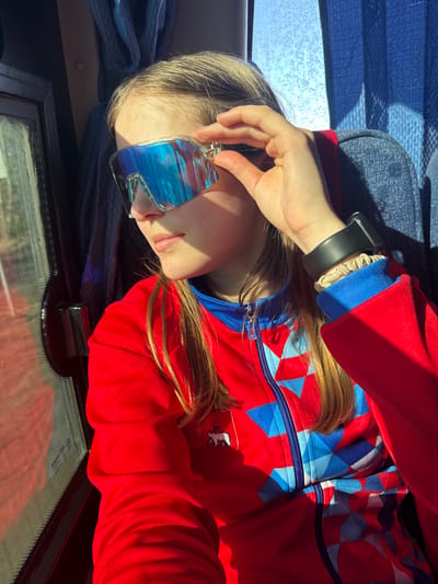 A young person with blond hair is resting in a seated position, possibly in a vehicle. They are wearing a red and blue patterned jacket and mirrored sunglasses. The person has a black smartwatch on their left wrist. The location is unclear, and there is no definitive evidence to confirm the scene is in the city of Челва, Russia. There are reflections in the window of what appears to be some vegetation.