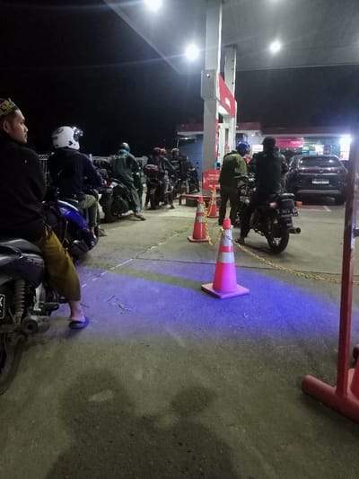 The image captures a nighttime scene at a gas station in Cimaung, Indonesia, bustling with activity. The primary subjects are numerous people, mostly on motorcycles, appearing to queue for fuel. The gas station is well-lit by overhead artificial lights, illuminating the area and casting distinct shadows. A prominent purple-blue light is cast on the ground in the foreground, possibly from a decorative light source or reflection.

In the left foreground, a man wearing a dark hoodie and a traditional cap (kufi or peci) sits on a blue and silver motorcycle, looking away from the camera. He is barefoot, wearing flip-flops. A line of motorcyclists, many wearing helmets, extends behind him towards the gas pumps, indicating a busy period. The gas station canopy and pumps display red and white accents, typical of Pertamina, Indonesia's national oil company.

Orange traffic cones with reflective white bands, linked by yellow chains, are positioned to direct vehicle flow. One cone in the center foreground is particularly bright due to the blue light. In the background, a dark car with the license plate "D 3868 TOP" is visible near the pumps. Another motorcycle in the right foreground has a license plate reading "D 3613 BM". A red sign near the pumps appears to have text, possibly a religious or safety message, which includes "PERHATIAN ALLAH S.W.T." The scene conveys a sense of routine evening or night activity at a gas station.