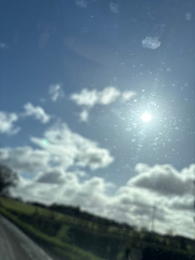 The image depicts an outdoor daytime scene captured through a significantly smudged and dusty transparent pane, likely a vehicle window. The location is Ruyton-XI-Towns, United Kingdom. The upper two-thirds of the frame show a blue sky with scattered white clouds. The sun, a bright spherical light source, is positioned in the upper right quadrant, creating lens flare and illuminating numerous sharp, irregular smudges, dust particles, and streaks on the glass surface. The lower third of the image reveals a highly blurred rural landscape, consisting of green fields or hills sloping upwards from left to right, interspersed with indistinct dark vegetation such as trees or hedges. A dark, diagonal element is visible along the bottom left edge, consistent with a road surface or vehicle interior component. No individuals are present, and no discernible actions or interactions occur within the external environment.