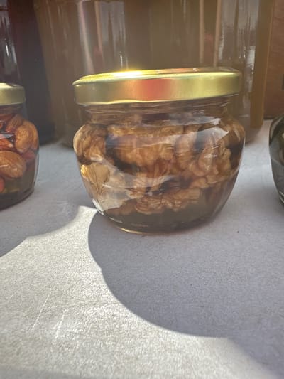 In the city of Усмангали, Russia, an image displays a close-up of a glass jar filled with walnuts and honey. The jar has a gold-colored lid. The jar is placed on a textured, light-colored surface, likely a table, and is illuminated by natural light, casting a shadow to the left of the jar. In the background, out of focus, are other glass containers with dark contents. To the left of the main jar, another similar jar is visible, containing almonds in honey.