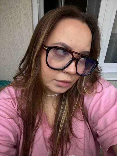 A woman with brown hair, wearing eyeglasses and a pink shirt, is visible in the image. She has a nose ring and is wearing a white beaded necklace. The background shows a textured wall and a window frame. The location is potentially Prigorodny, Russia.