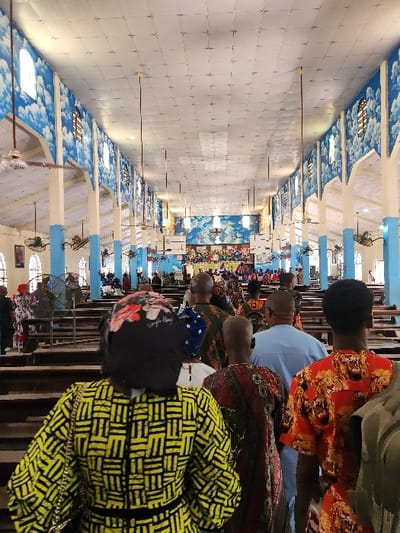 This image depicts a large indoor gathering, likely a religious service or communal event, in a spacious hall or church in Umuoye, Nigeria. The perspective is from the back of the hall, looking towards the front, with rows of people seated in pews.
The subjects are numerous individuals, primarily adults, seated on wooden benches or pews. Many are seen from behind, showing the backs of their heads and shoulders. Their attire is varied, featuring colorful patterns; for example, one person in the foreground wears a bright yellow and black geometric patterned garment, and another has an orange floral pattern. Several people wear head coverings or scarves.
The setting is a long hall with a high ceiling. The interior decor is distinctive: the ceiling and upper walls are adorned with murals depicting blue sky and white clouds, creating an expansive, open feel. Light-colored pillars or columns, some painted with light blue sections, line the hall. Multiple ceiling fans are visible, suggesting a need for ventilation. The floor is covered with light-colored tiles, arranged in a grid pattern. In the distance, at the far end of the hall, there appears to be an altar or stage area with more vibrant decorations and potentially another mural or artwork. The overall arrangement with pews indicates a structured gathering.
The activity appears to be a formal assembly, such as a church service or a community meeting, where attendees are seated and observing what is happening at the front of the hall. The orderly seating arrangement and the nature of the venue suggest a solemn or respectful atmosphere.
Notable details include the unique sky-and-cloud ceiling design, which could be symbolic in a religious context. The time of day appears to be daytime, as the interior is well-lit, although there are no direct external views to confirm this. No discernible text is visible anywhere in the image. The vibrant clothing patterns are characteristic of attire often seen in West African communities.