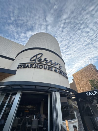 The image displays the exterior of "Perry's Steakhouse & Grill" under a bright, cloudy sky in Oak Brook, United States of America. The restaurant building features a distinctive curved facade in a light, off-white color, with a subtle textured pattern. The prominent sign, "Perry's Steakhouse & Grill," is rendered in dark, elegant script and block letters on this curved surface.

To the right of the steakhouse, another building with a textured, rough brown stone facade is visible, rising against the sky. In the immediate foreground on the right side of the image, a black structure, possibly an awning or part of an entrance canopy, prominently displays the word "VALET" in white capital letters. Below this, large glass windows or doors offer reflections of the interior and surrounding environment.

The sky is filled with numerous small, scattered white clouds, suggesting a partly cloudy day. There are no discernible people or specific activities taking place, and the scene is primarily an architectural shot of the restaurant's exterior. The visible text clearly identifies the establishment and indicates the presence of a valet service.