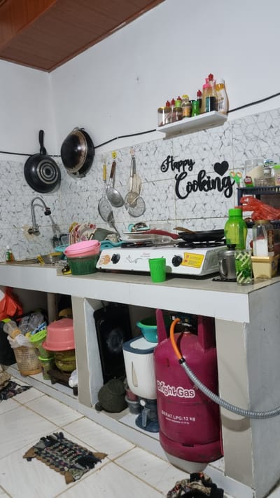 The image shows a cluttered kitchen counter in Batam, Indonesia. The counter is made of white laminate and is filled with various kitchen items, including pots, pans, utensils, dishes, and cooking appliances. A white gas stove is positioned on the counter, with a black frying pan placed on top of it. To the right of the stove, there is a pink gas cylinder labeled "Bright Gas" and "LPG-12 kg". Several plastic containers and baskets are also visible on the counter and in the shelves below. The wall behind the counter is tiled with a geometric pattern, and a shelf above it holds an assortment of spice bottles and other condiments. The overall impression is of a functional, albeit somewhat disorganized, kitchen space. The flooring appears to be white tiles, and a small woven mat is placed on the floor in the foreground.