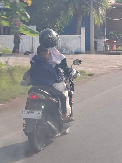 In Keudealuerheung, Indonesia, a man and a child are riding a dark-colored motorcycle. The man is in the front, wearing a dark hoodie and black pants, with a backpack slung over his shoulder. The child is behind him, wearing a light-colored headscarf and what appears to be a pinkish garment. The motorcycle has a license plate that reads "BL 3699 ZBF". The scene is outdoors, with a paved road and grassy areas visible. In the background, there are buildings, palm trees, and a person sweeping with a broom. The lighting suggests it is daytime, possibly late afternoon due to the angle of the sun and the shadows cast.