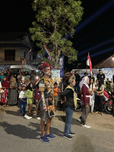 This image depicts a nighttime public gathering or event in Kedungbanteng, Indonesia. The foreground features an asphalt road surface with shadows cast by artificial lighting. Centrally, a large tree with green foliage stands against the dark sky.

In the midground, a crowd of individuals is present. Three figures are prominent: on the left, a male wearing a feathered headdress with red and black plumes, white face paint, a patterned black, orange, and yellow tunic over a dark shirt, grey shorts, and blue athletic shoes; to his immediate right, a female wearing a feathered headdress, a dark long-sleeved top, blue jeans, and black slip-on shoes; further right, a female wearing a white hijab, a maroon long-sleeved top, a patterned dark skirt, and white athletic shoes, with her mouth slightly open, suggesting speech or song.

The background contains numerous onlookers, including adults and children, some wearing head coverings. Structures include a two-story building on the left with a dark roof and light-colored walls, and a building with a thatched roof on the right. Banners and posters are visible, one displaying "DESA KA..." and another advertising "LAS AUDIO." Multiple red and white Indonesian flags are affixed to poles or structures. Beams of light from external spotlights project into the dark night sky from the right. A red and white scooter is parked on the far right side of the frame.