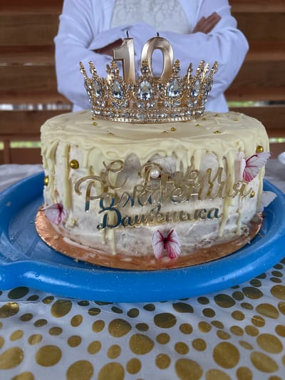 A 10th birthday cake with a gold crown and "Happy Birthday Daughter" written in gold lettering is displayed on a blue tray. The cake is white with white frosting dripping down the sides and decorated with gold beads and pink butterflies. A person wearing a white top stands in the background, with their arms crossed. The scene appears to be set outdoors, possibly at a celebration in Ганчарскі сельскі Савет, Belarus, with a wooden structure visible in the background and a gold-spotted tablecloth in the foreground.